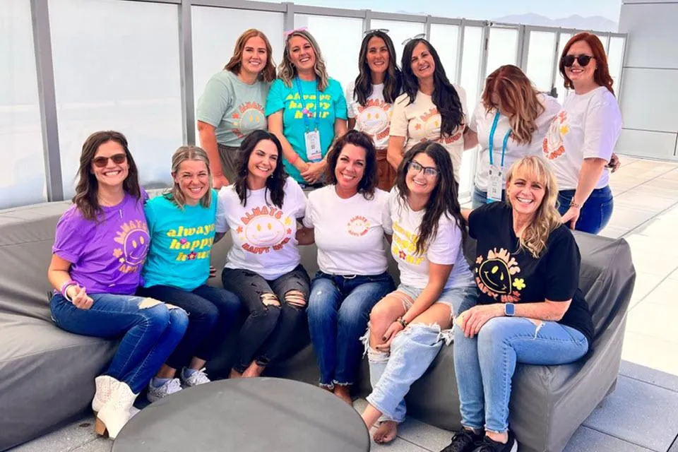 A group of twelve smiling women pose together on a rooftop lounge with a bright blue sky and mountains in the background. They are wearing colorful "Always Happy Hour" branded t-shirts in various shades, paired with jeans and casual footwear. Some are sitting on a gray outdoor couch while others stand behind them, radiating positivity and team spirit in a relaxed, modern setting.