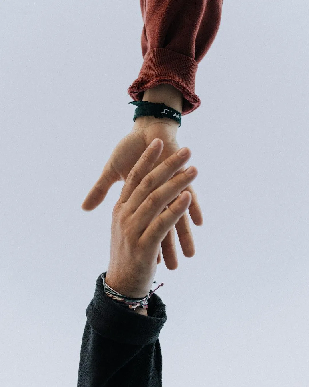 Two hands reaching for each other against a light, neutral background. One hand, wearing a red long-sleeve sweater and a green wristband, extends downward, while the other, adorned with multiple string bracelets and a black sleeve, reaches up. The image symbolizes connection, support, and helping hands.