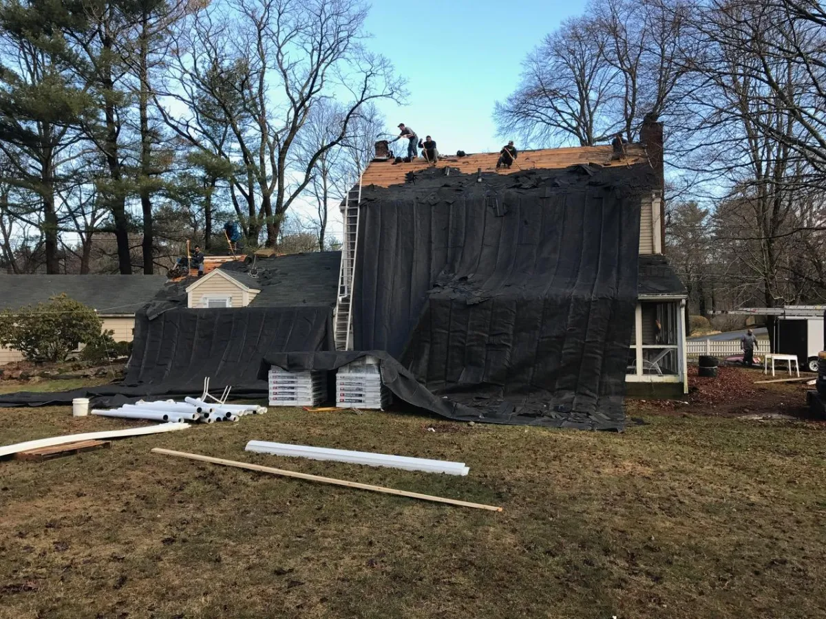 Roof replacement in progress by McCarthy Exteriors LLC on a residential home in Brookline, NH with protective tarp and crew at work