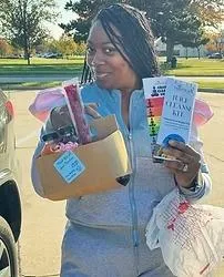 A woman in a gray tracksuit holds a cardboard box with various items and a flu clean kit pamphlet while standing outdoors.