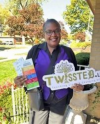A woman in glasses and a purple and black outfit stands outside, holding brochures and a "TWISSTED" sign. Trees, grass, and houses are visible in the background.