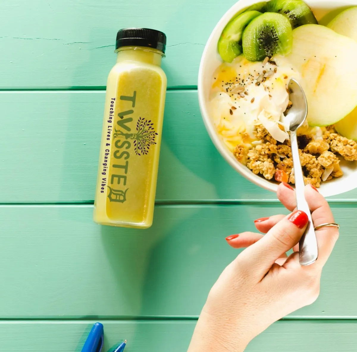 A hand holds a spoon over a yogurt bowl topped with fruit and granola, next to a bottle of yellow juice on a teal table.