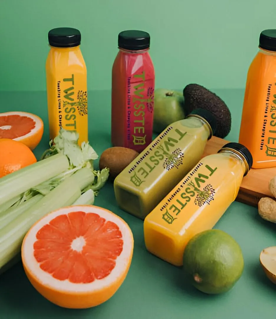Colorful juice bottles from "Twissted" surrounded by fresh fruits and vegetables on a green background. Fresh, vibrant, and healthy.