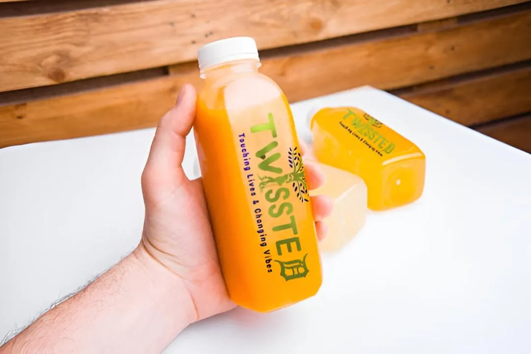 A hand holding a bottle of orange juice labeled "TWISSTED" with the tagline "Touching Lives & Changing Vibes," with other juice containers in the background.