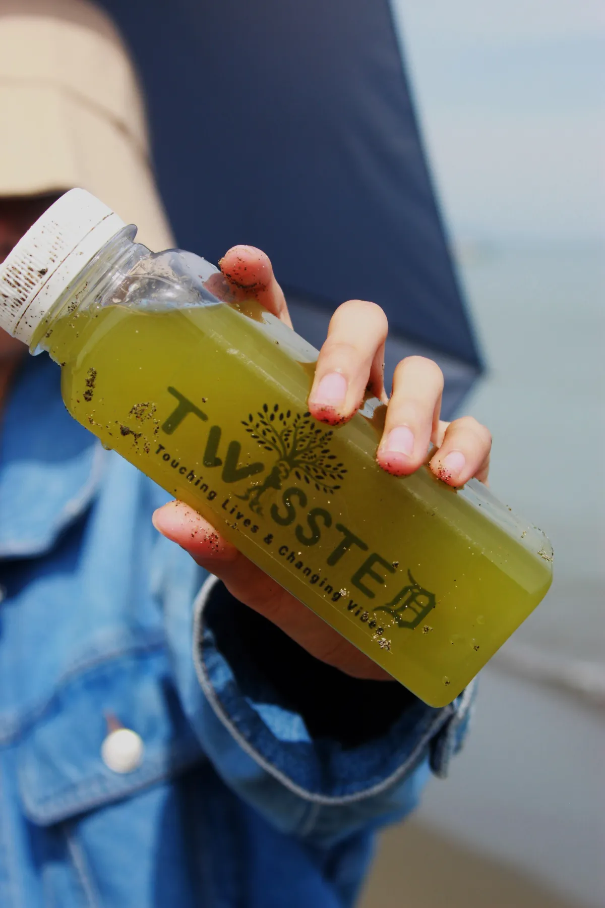 Person in a denim jacket holding a green TWISSTED juice bottle with sand on the fingers and bottle, set against a beach background. Keywords: TWISSTED green juice, healthy beach lifestyle, cold-pressed wellness drink, detox beverage, natural juice brand photography, organic nutrition drink.