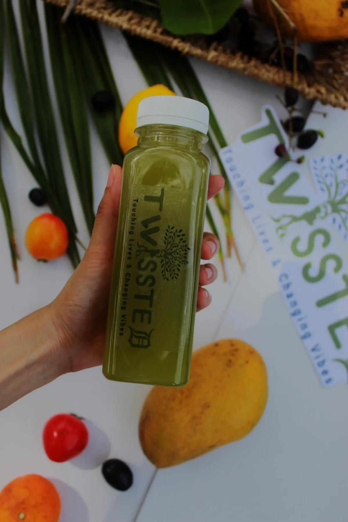 Hand holding a green TWISSTED juice bottle with the logo “Touching Lives & Changing Vibes,” surrounded by fresh fruits and vegetables—tomatoes, mango, blueberries, and palm leaves—on a white background. Keywords: green detox juice, TWISSTED health drink, plant-based beverage, cold-pressed juice photography, wellness product branding, clean eating concept.