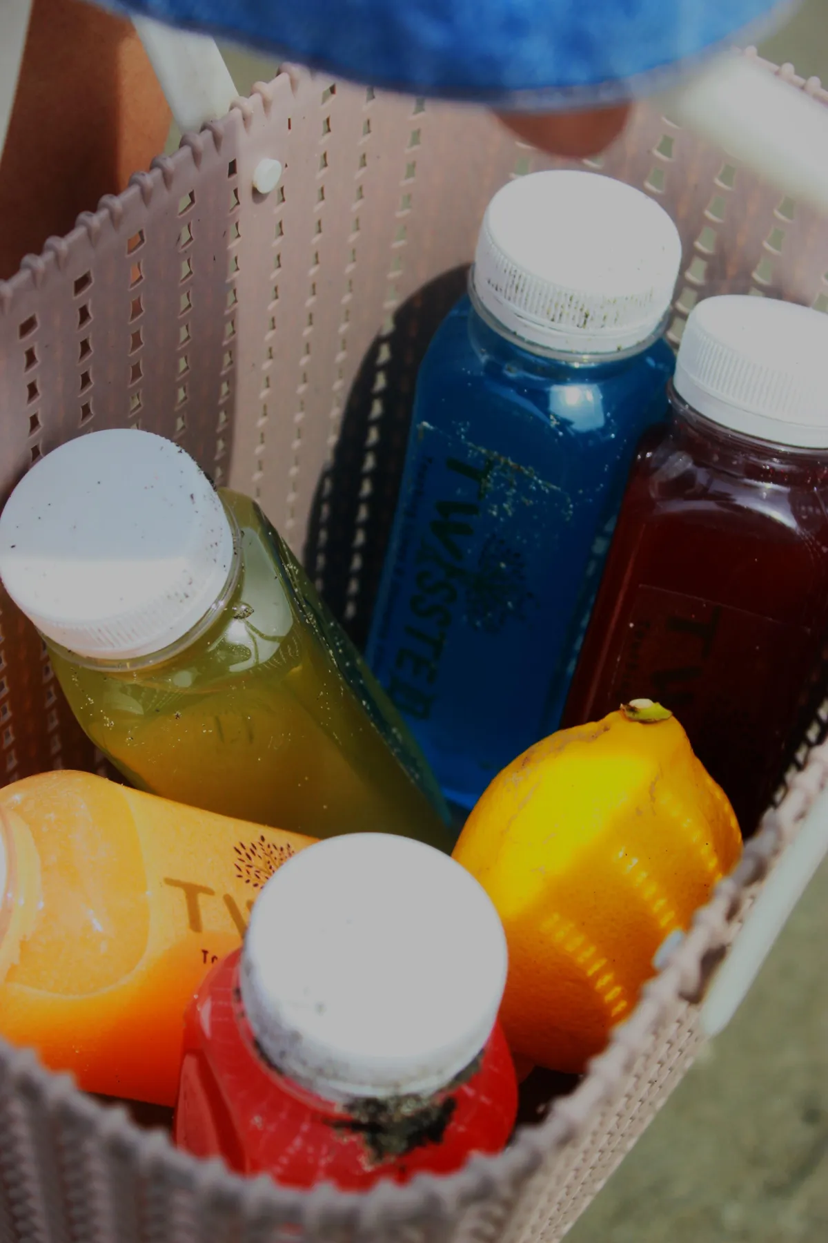 Overhead view of a woven basket containing TWISSTED bottled juices in red, blue, orange, and green, along with a fresh lemon. The bottles are partially covered in sand, suggesting a beach outing. Keywords: TWYSSTE detox juices, beach picnic drinks, healthy lifestyle beverages, fruit-based cold-pressed juices, vitamin-rich juice bottles.