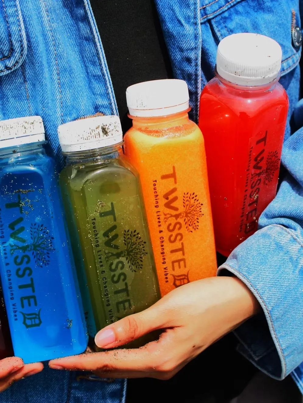 Close-up of a person holding five colorful TWISSTED juice bottles—purple, blue, green, orange, and red—against a denim jacket. The bottles are labeled “Touching Lives & Changing Vibes.” Keywords: TWISSTED juice collection, cold-pressed juice, colorful detox drinks, wellness lifestyle, healthy beverage branding, natural fruit juice, multicolor juice bottles.