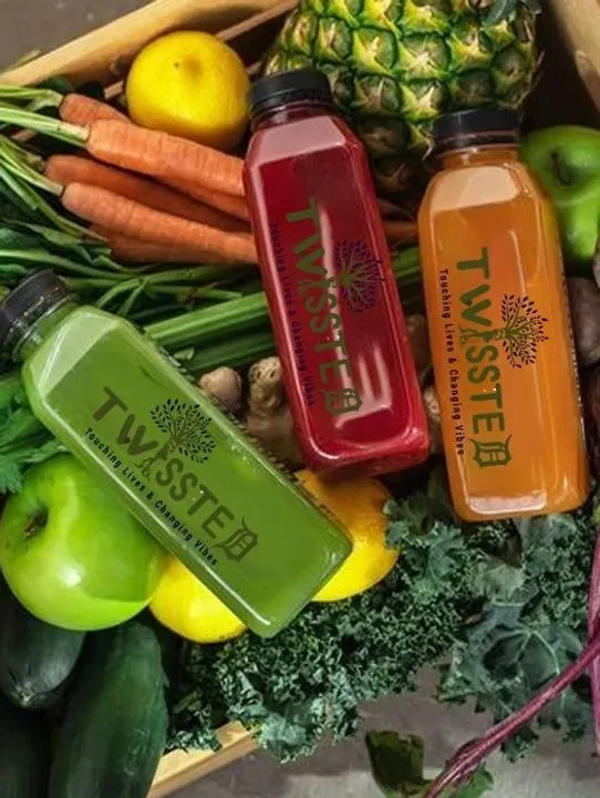 Three colorful TWISSTED cold-pressed juice bottles—green, red, and orange—displayed on a bed of fresh fruits and vegetables including pineapple, carrots, apples, kale, lemons, and cucumbers. Each bottle features the brand logo “TWISSTED – Touching Lives & Changing Vibes.” Keywords: TWISSTED juice, cold-pressed detox drinks, fruit and vegetable juice, healthy lifestyle beverage, organic juice cleanse, wellness drink photography.