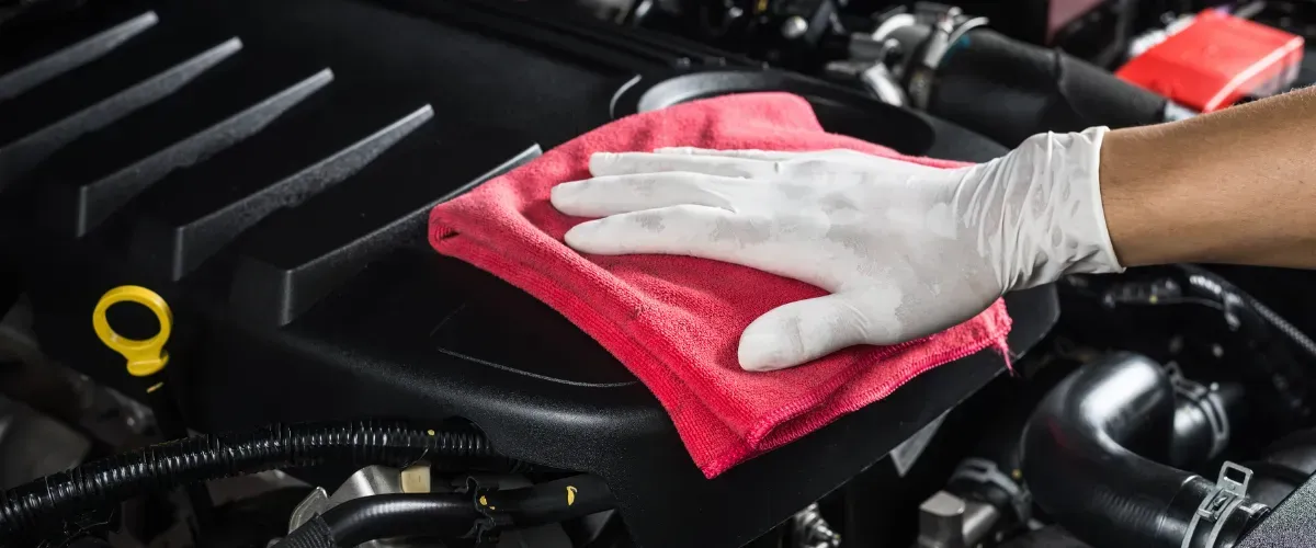 Gloved hand cleaning a car engine bay with a red microfiber cloth