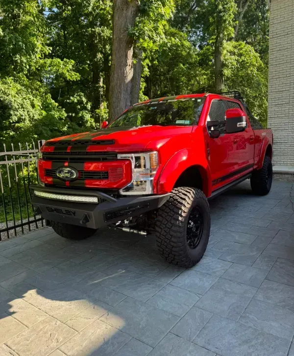 Red lifted Ford Shelby truck parked on a stone driveway near a forested area.