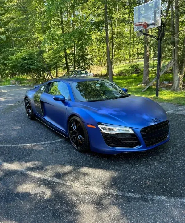 Blue Audi R8 sports car parked on a residential driveway surrounded by trees on a sunny day.