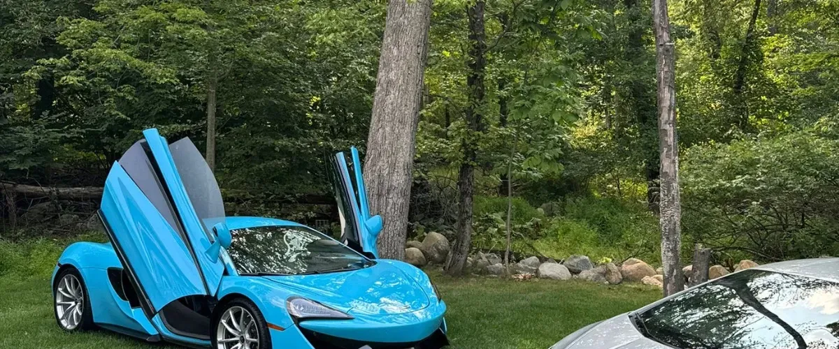 Blue McLaren supercar with butterfly doors open, parked on grass near a wooded area.