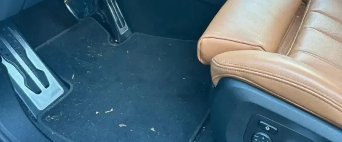 Dirty car floor mat with debris near pedals and brown leather seat before interior detailing