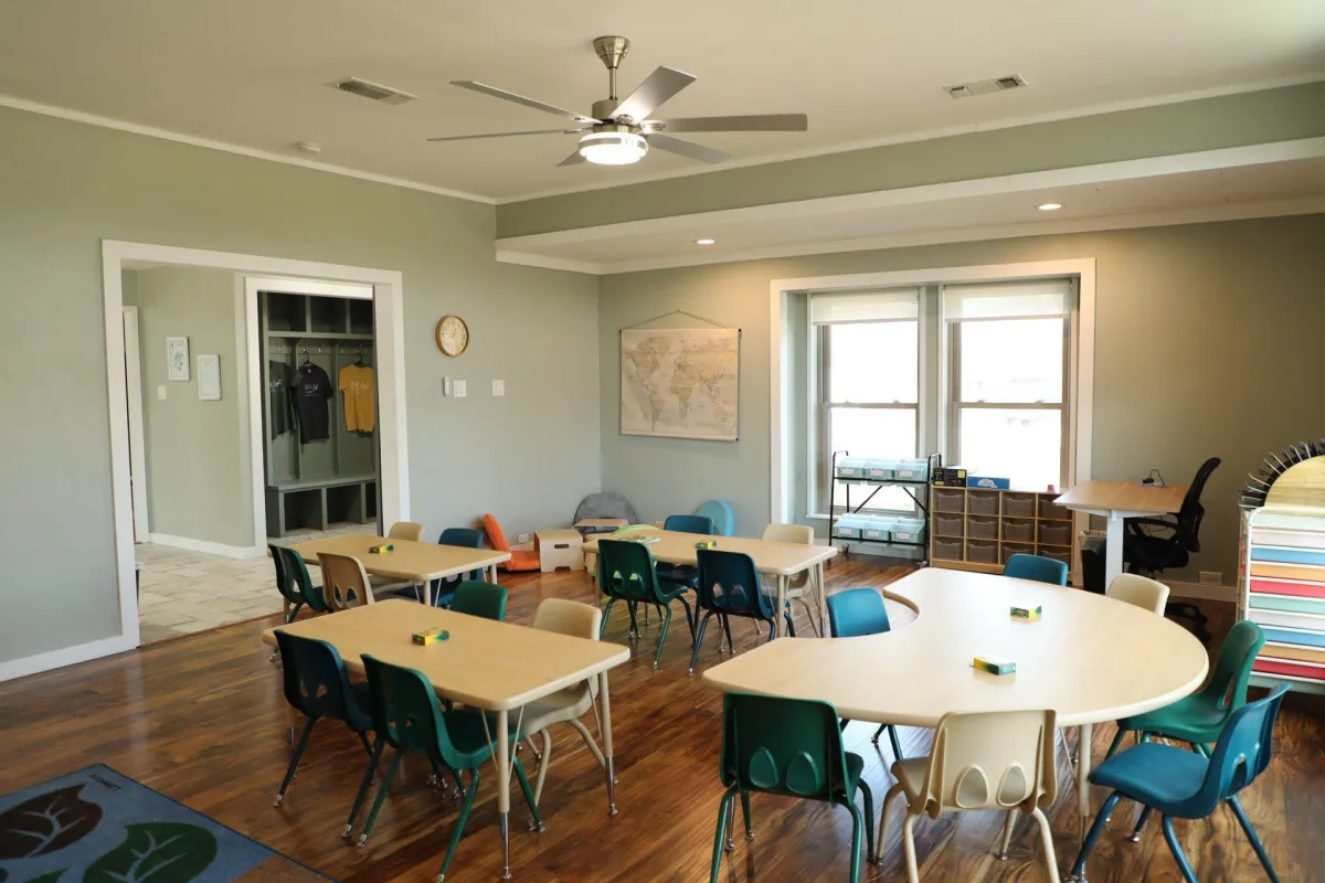Children tending a vibrant vegetable garden, guided by a teacher in a straw hat, with a backdrop of a white picket fence and distant chapel. The scene is lively, educational, and faith-inspired.