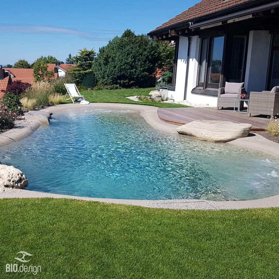 Ein naturnah gestalteter Pool mit klarem, türkisblauem Wasser, umgeben von einer sanft geschwungenen Uferzone mit Sandoptik und Kieselsteinen. Eine Holzterrasse mit bequemen Gartenmöbeln grenzt an das Haus mit großen Fenstern. Im Hintergrund befinden sich eine grüne Rasenfläche, Zierpflanzen und ein weißer Liegestuhl. Das gesamte Ambiente wirkt harmonisch und natürlich, passend zur umgebenden Landschaft mit Bäumen und roten Ziegeldächern.