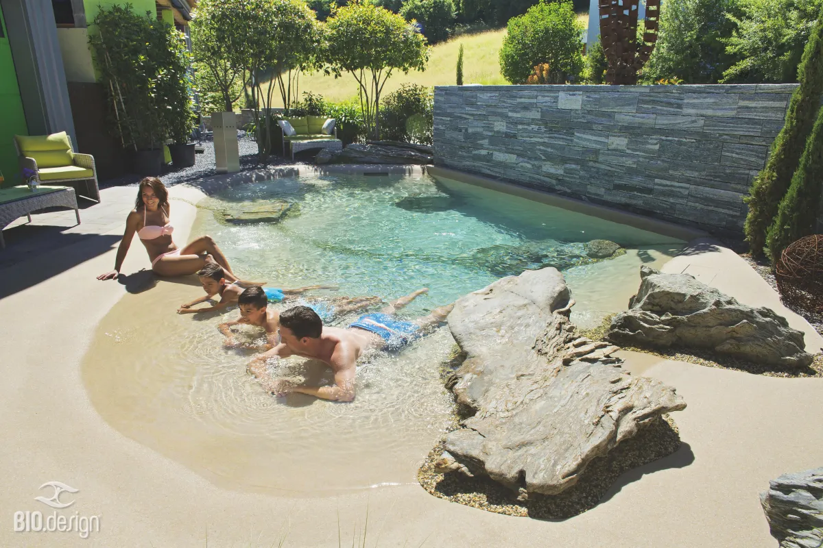 Familie genießt den flachen Bereich eines Naturpools mit sandigem Ufer und natürlicher Steinlandschaft. Die Kinder spielen im seichten Wasser, während die Mutter entspannt am Beckenrand sitzt. Der Pool ist umgeben von modernen Gartenmöbeln und einer stilvollen Steinmauer.