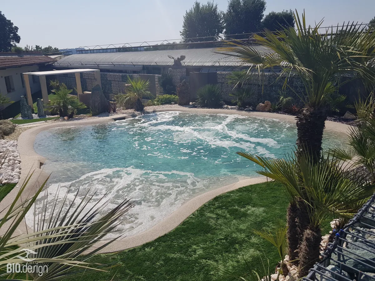 Ein natürlicher, lagunenartiger Pool mit türkisblauem Wasser und sanften Wellenbewegungen, umgeben von einer sandfarbenen Uferzone und tropischer Bepflanzung mit Palmen. Im Hintergrund befinden sich eine Steinmauer, bepflanzte Bereiche mit dekorativen Felsen sowie ein Gebäude mit überdachter Terrasse. Die Umgebung vermittelt ein mediterranes Ambiente mit gepflegtem Grün und weißen Kieselsteinen.