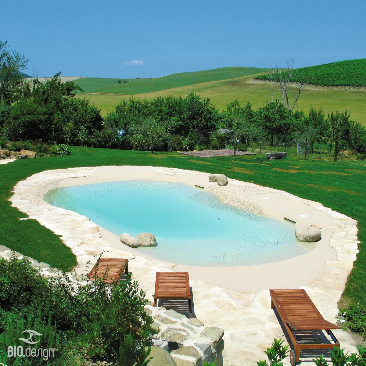 Ein natürlicher, lagunenartiger Pool mit hellblauem Wasser, umgeben von einer sandfarbenen und steinernen Uferzone, eingebettet in eine weitläufige grüne Landschaft mit sanften Hügeln im Hintergrund. Mehrere Holzliegen stehen am Rand des Pools auf der steinernen Fläche. Die Umgebung ist geprägt von gepflegtem Rasen, Bäumen und Sträuchern, die eine mediterrane und naturnahe Atmosphäre schaffen.