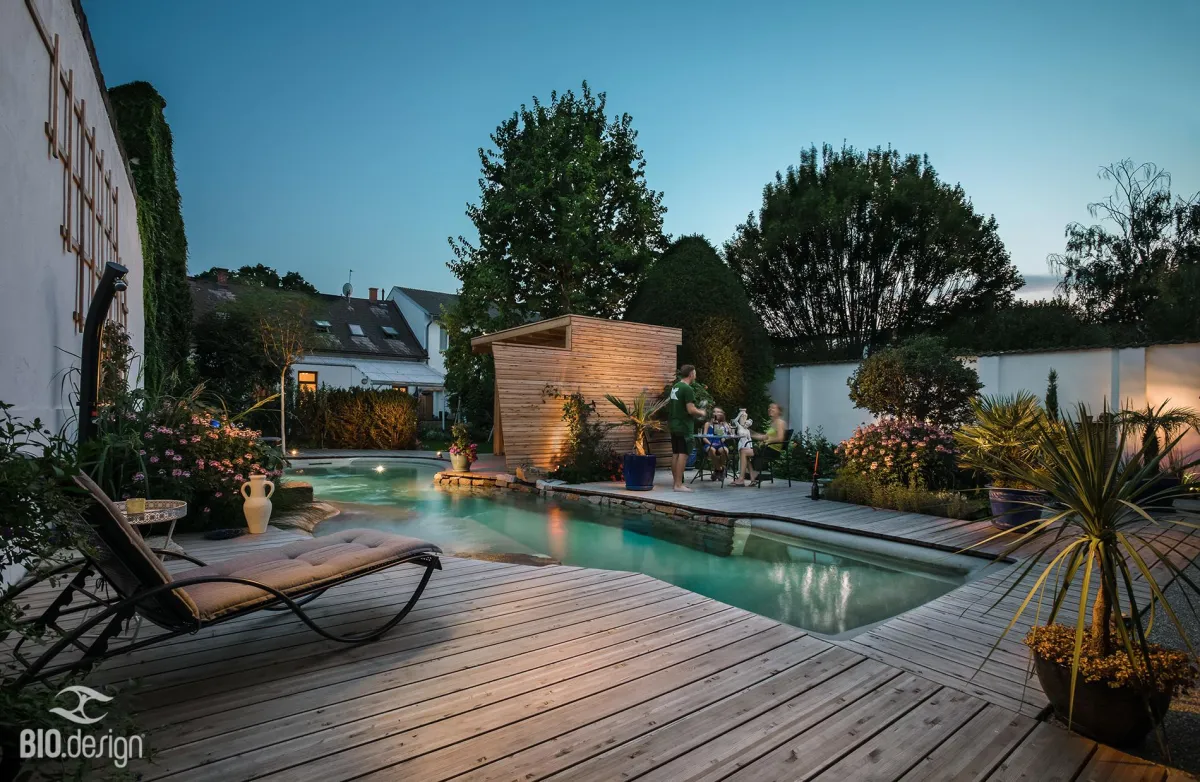 Ein stilvoll gestalteter Naturpool in einem modernen Garten bei Abenddämmerung. Die Poollandschaft ist von einer großzügigen Holzterrasse, blühenden Pflanzen und dekorativen Beleuchtungen umgeben. Eine Sonnenliege steht im Vordergrund auf der Terrasse, während im Hintergrund eine Gruppe von Menschen an einem Tisch sitzt und eine gemütliche Atmosphäre genießt. Ein kleines Holzhaus mit indirekter Beleuchtung ergänzt das harmonische Ambiente. Die sanfte Poolbeleuchtung spiegelt sich im klaren Wasser wider, wodurch eine entspannte und einladende Stimmung entsteht.