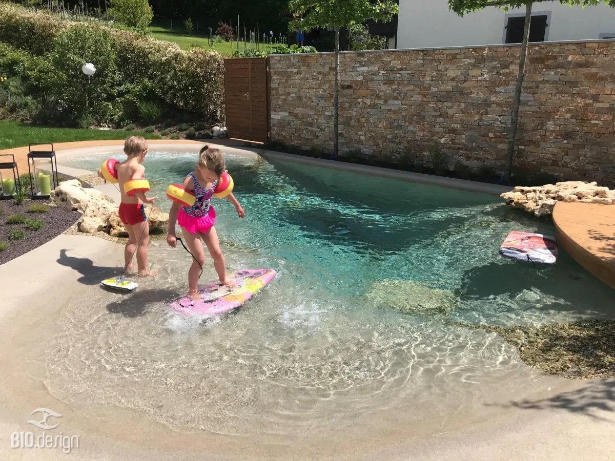 Zwei Kinder mit Schwimmflügeln spielen in einem flachen, lagunenartigen Pool mit natürlicher Sandoptik. Sie stehen auf kleinen Bodyboards und lassen sich vom Wasser tragen. Der Pool ist von einer geschwungenen Uferzone mit Steinen, Holzdecks und einer dekorativen Steinmauer umgeben. Im Hintergrund wachsen Bäume und Pflanzen, die das mediterrane Ambiente des Gartens unterstreichen.