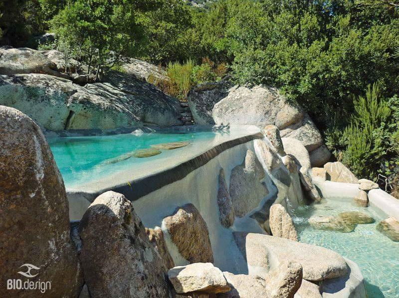 Ein beeindruckender, naturnaher Infinity-Pool, der sich harmonisch in eine felsige Landschaft einfügt. Das türkisfarbene Wasser fließt sanft über eine natürliche Kante in ein darunter liegendes Becken, das von großen, abgerundeten Felsen umgeben ist. Die Umgebung ist dicht mit grüner Vegetation bewachsen, wodurch der Pool wie ein natürlicher Gebirgsbach wirkt. Der Poolbereich vermittelt eine luxuriöse, aber dennoch wilde und organische Atmosphäre, perfekt eingebettet in die Natur.