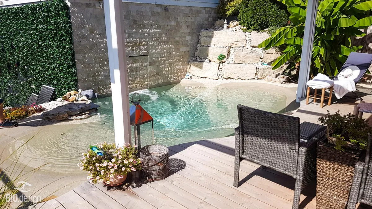 Ein stilvoll gestalteter Naturpool mit klarem Wasser und einer flachen, sandfarbenen Uferzone, integriert in eine gemütliche Gartenoase. Im Hintergrund fließt Wasser aus einer modernen Natursteinmauer in den Pool. Eine erhöhte Terrasse mit großen Natursteinen und tropischen Pflanzen, darunter eine Bananenstaude, sorgt für eine entspannte Atmosphäre. Im Vordergrund stehen Gartenmöbel aus Rattan auf einer Holzterrasse, dekoriert mit Blumen, Laternen und Accessoires, während eine Sonnenliege mit Handtüchern zum Entspannen einlädt.