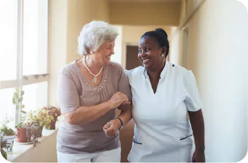 An elderly white woman and a black female nurse walking hand in glove and laughing