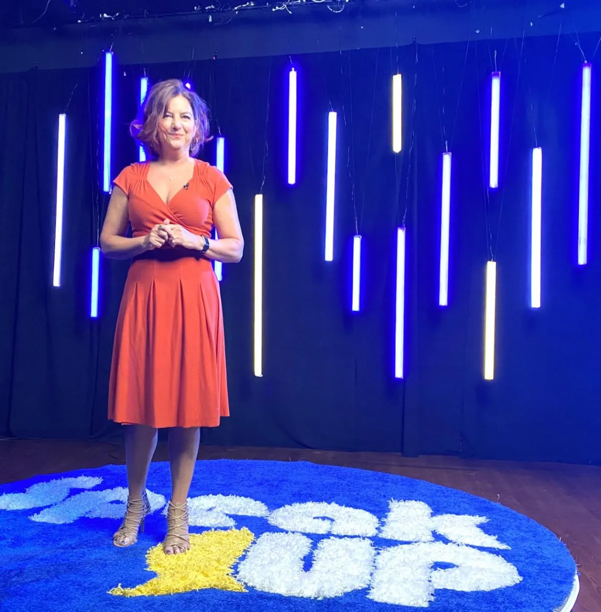 A woman stands on stage in front of a backdrop with vertical blue and white LED lights. She wears a red dress and beige heels, and her hands are clasped. The stage floor features the words "Speak Up" in blue and white lettering.