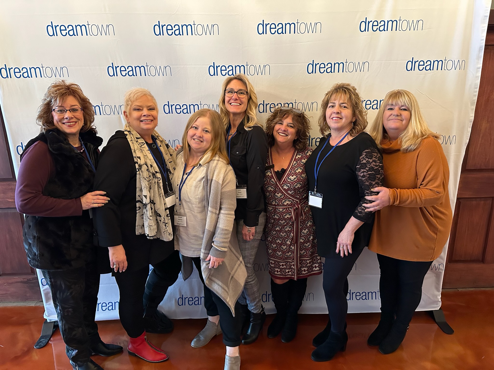 Seven women stand smiling in front of a "dreamtown" step and repeat backdrop. They are dressed in a variety of casual and semi-formal attire. Some wear name badges around their necks. The floor is a reddish-brown tile, and the backdrop is white with blue text.