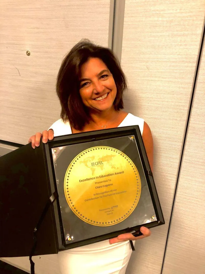 Clara Capano - A person with shoulder-length dark hair smiles while holding a gold plaque that reads "Excellence in Education Award" in English. The plaque is in a black case, and the person is wearing a white outfit. The background is an indoor setting with light-colored walls.