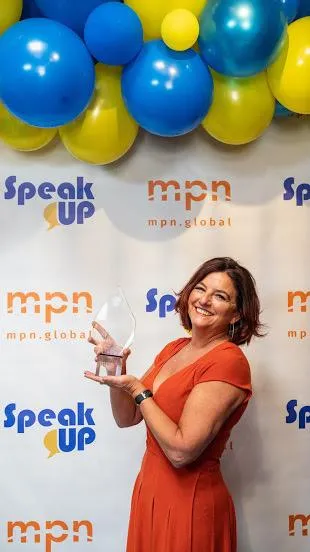 Clara Capano, a woman in an orange dress smiles while holding an award. She is standing in front of a backdrop with logos for "Speak Up" and "mpn.global." Blue and yellow balloons are clustered at the top of the backdrop.