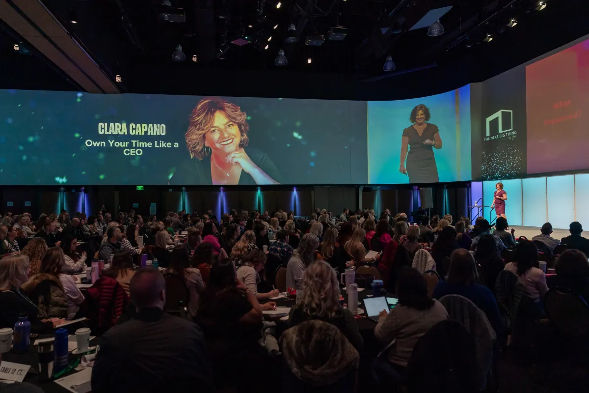 A woman with shoulder-length brown hair is speaking energetically on a stage. She is wearing a red dress and a microphone attached to her neckline. The text overlay reads "CLARA CAPANO" with subtext "International Speaker, Best Selling Author and Award Winning Educator.