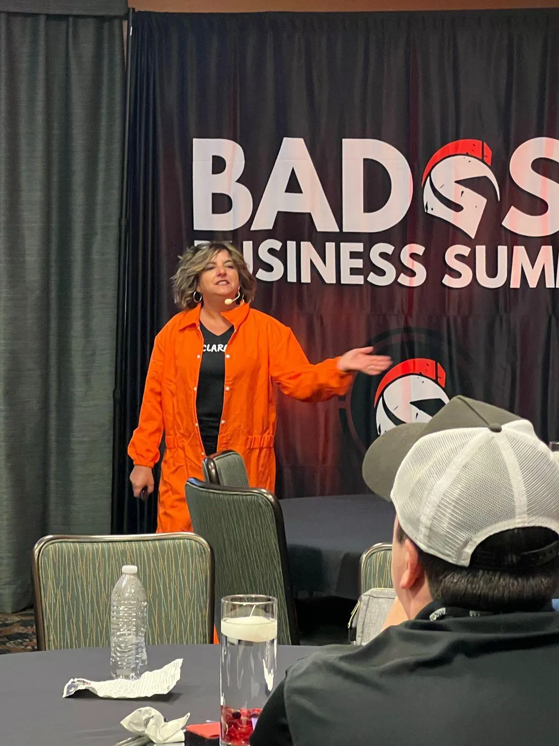 Clara Capano, dressed in an orange jumpsuit over a black shirt, is presenting on a stage at the "Badass Business Summit". The background features a black banner with the event name. The audience, seated at tables, includes a person in a white cap.