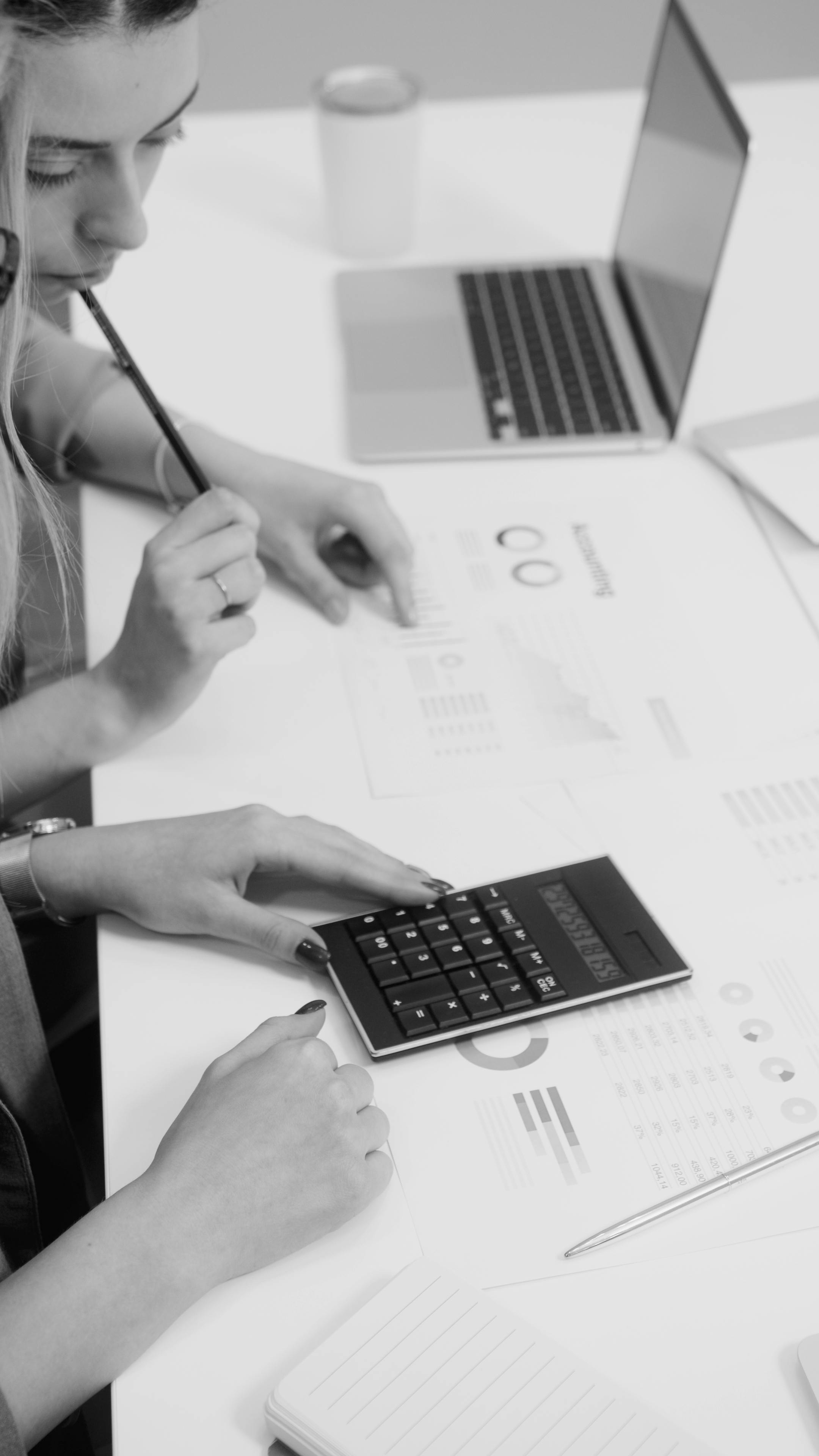Black and white photo of a professional analysing printed charts and using a calculator at a desk, reflecting the financial and performance impact of strategic decisions.