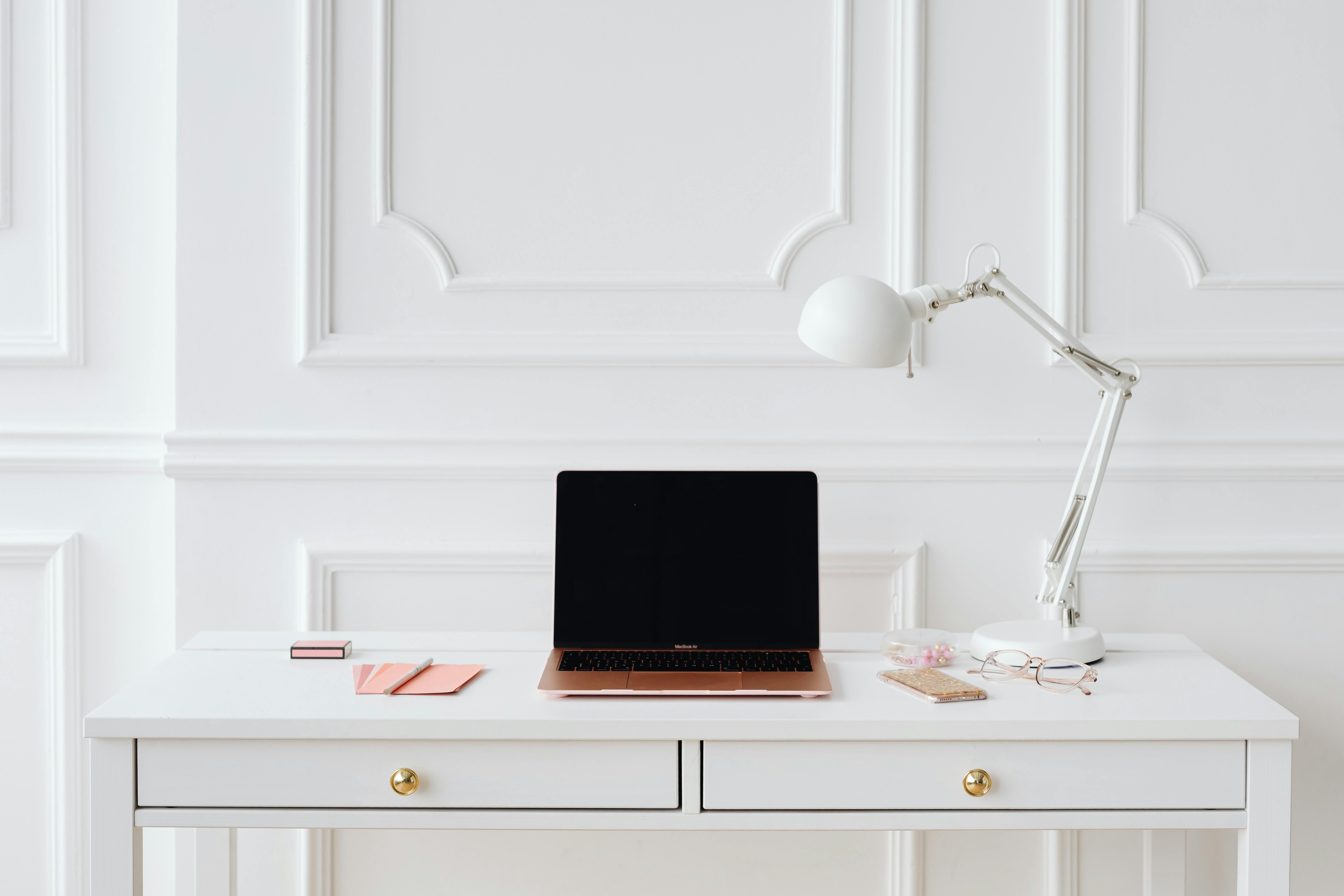 Clean white desk with rose gold laptop and lamp in minimalist office setting, representing structured and professional social media management.