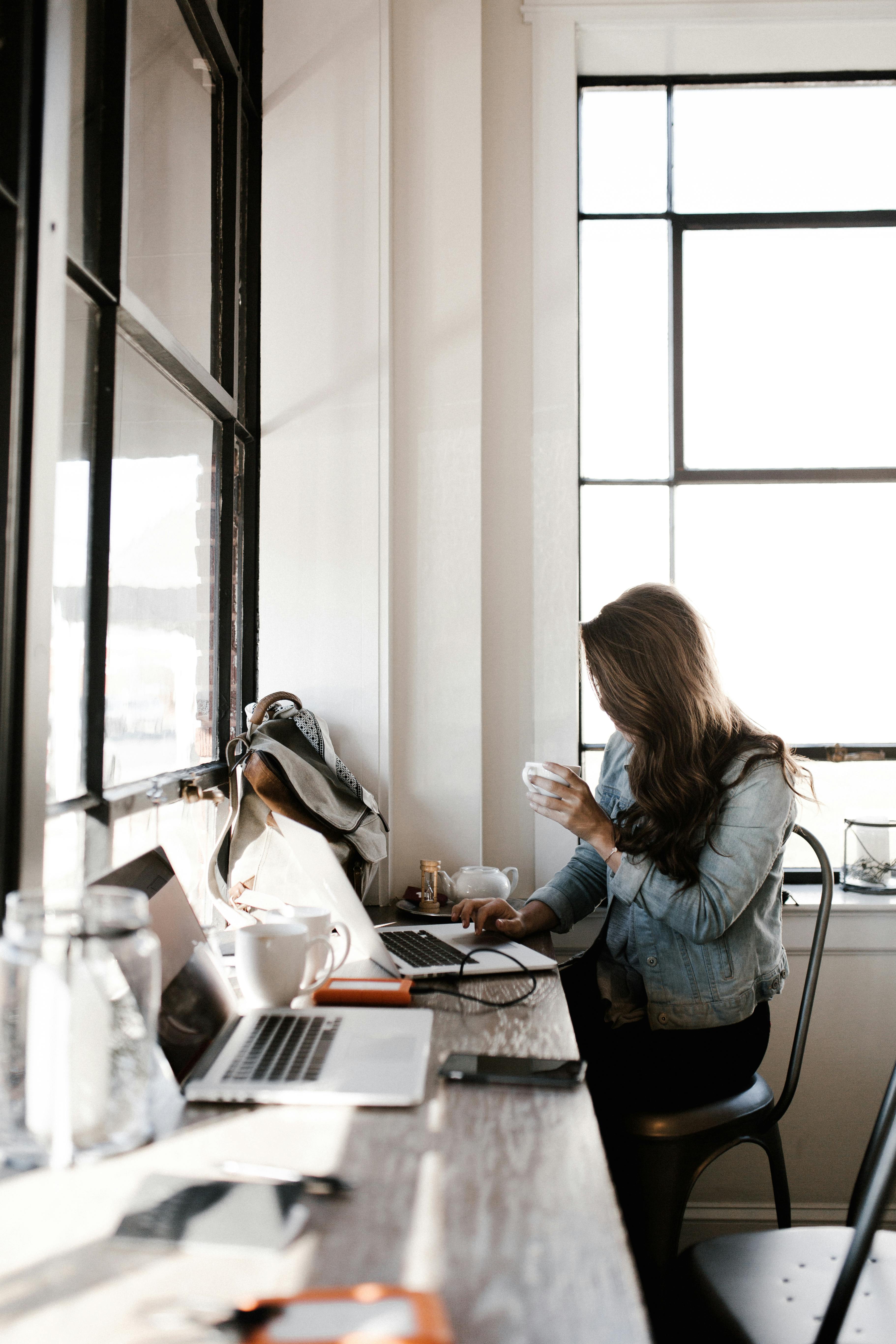 A woman sitting at a desk by large industrial windows, working on a laptop and holding a cup, viewed from behind in warm natural light.