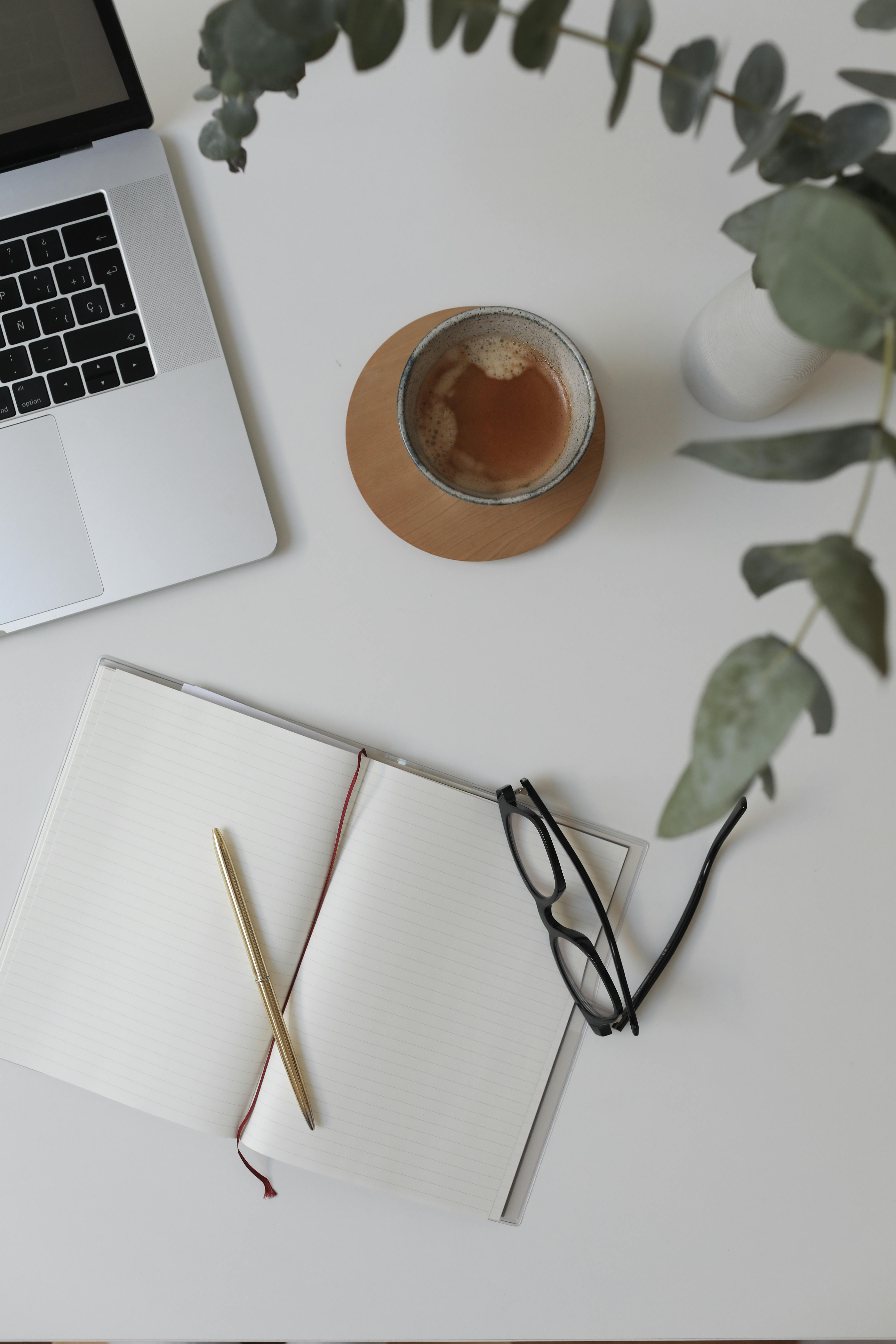 An overhead view of a clean white desk with an open notebook, a gold pen, a pair of glasses, a laptop and a cup of tea beside a eucalyptus plant.