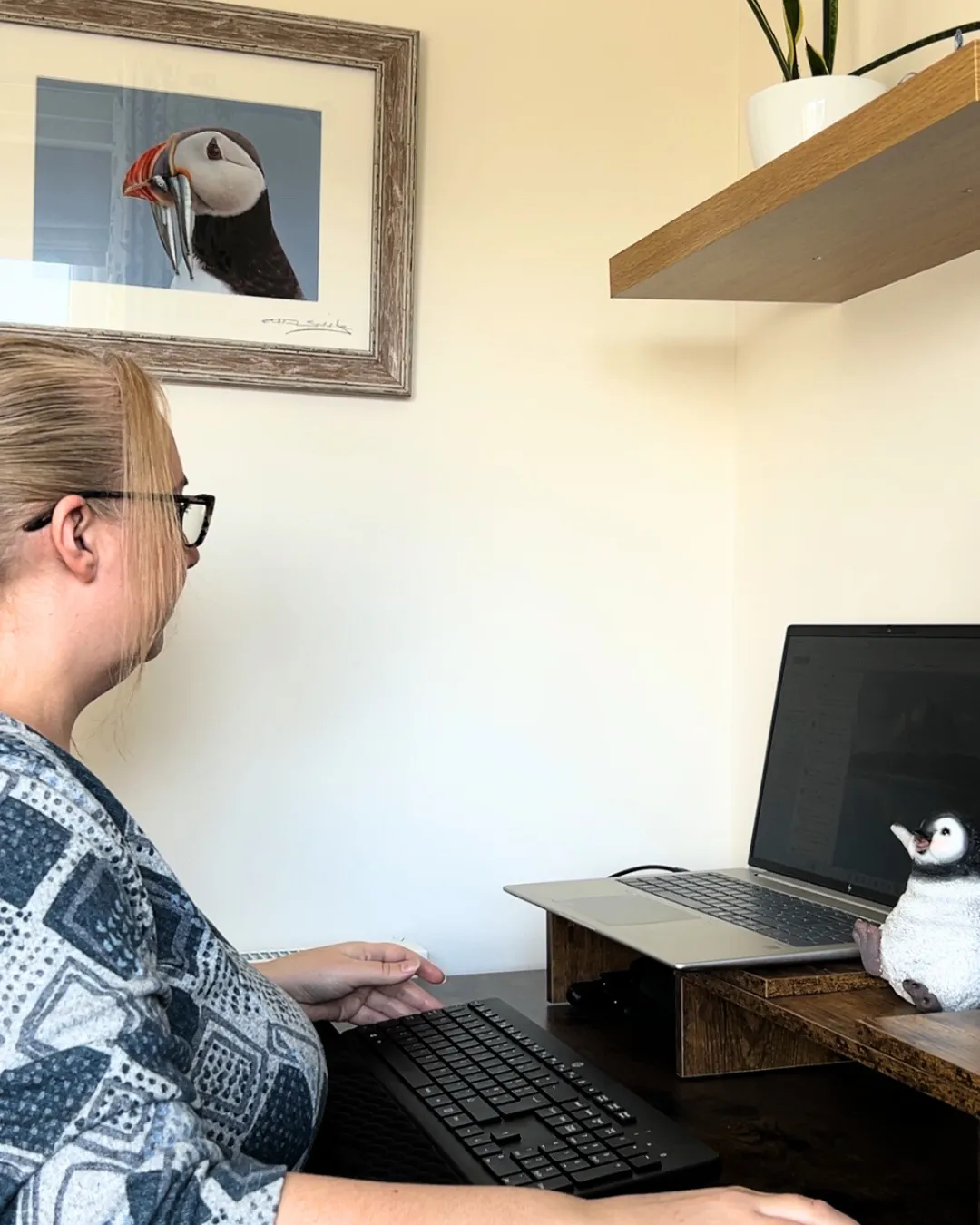 Hayley, founder of The Efficient Penguin Co., working at her desk with a small penguin ornament beside her laptop, a framed puffin print on the wall and a plant on the shelf above.