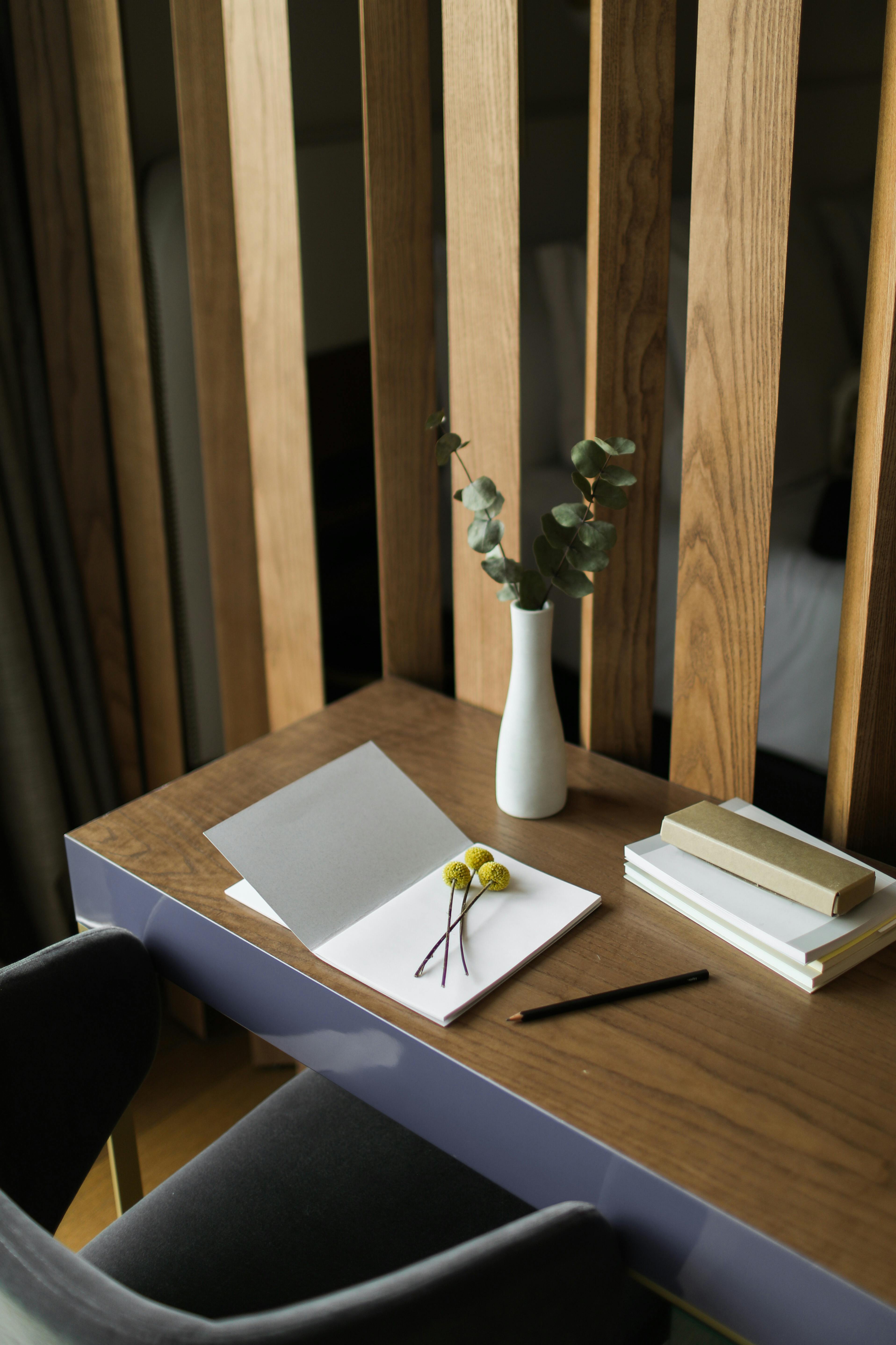 A warm wooden desk with an open document, a small notebook with dried yellow flowers, a stack of notebooks, a pencil and a white vase with eucalyptus, set against a wooden slatted backdrop.