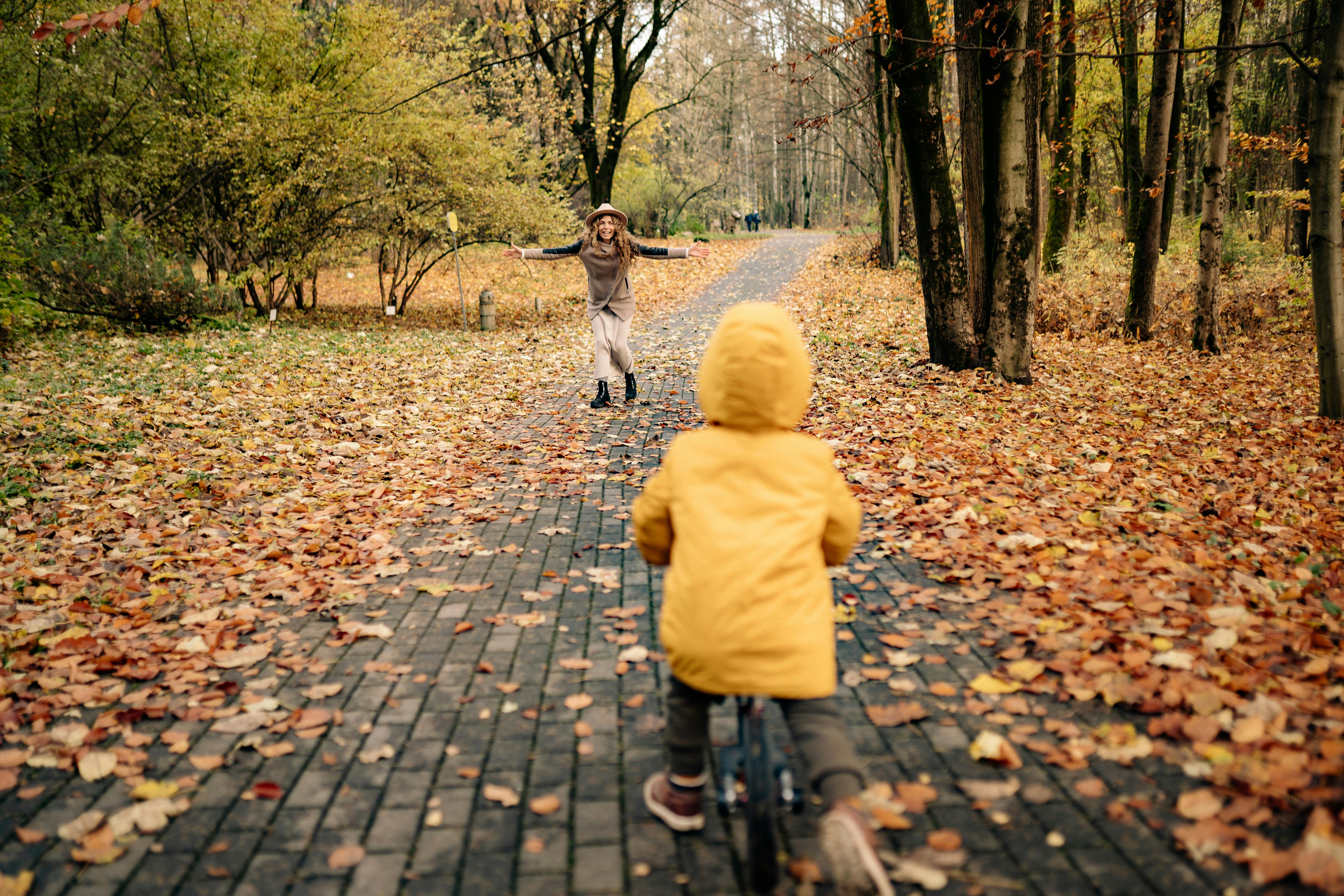A mother runs with arms open wide toward her young child on a bicycle along an autumn tree-lined path, capturing the feeling of being present and free from the weight of doing everything alone.