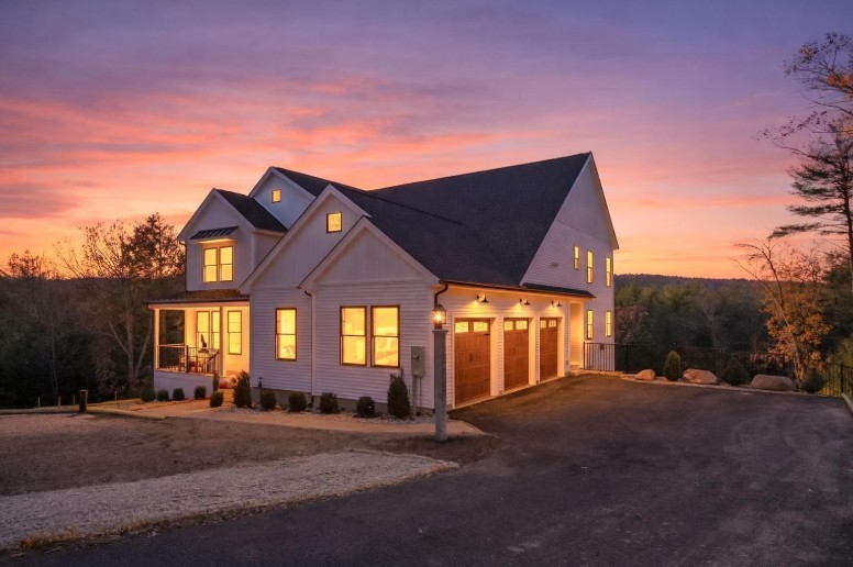 A modern white two-story home with a three-car wooden garage and glowing windows under a dramatic purple and orange sunset sky.