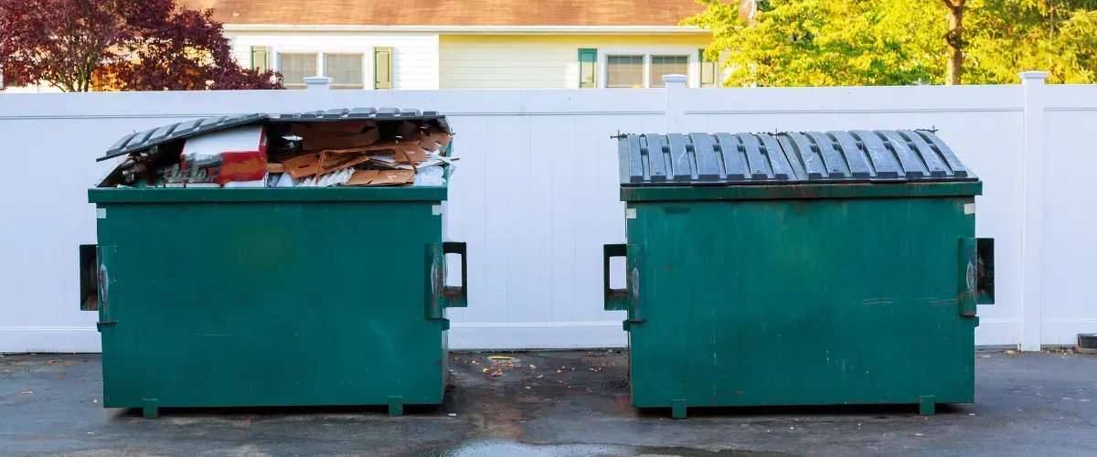 Two green dumpsters in a residential area, one overflowing with cardboard and trash against a white fence