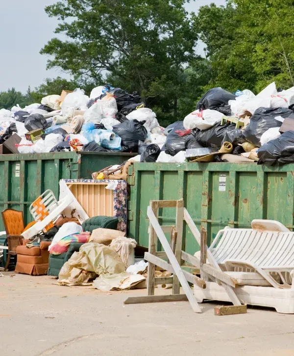 Large green dumpsters overflowing with black trash bags, furniture, and debris in an outdoor area