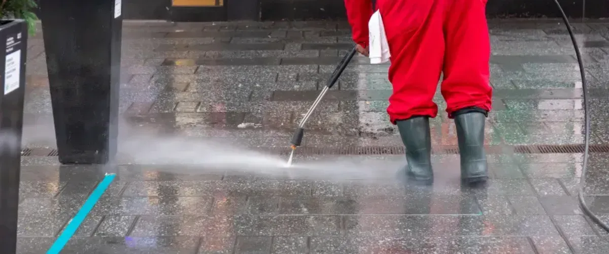Worker in red uniform pressure washing a wet city sidewalk to remove dirt and debris.