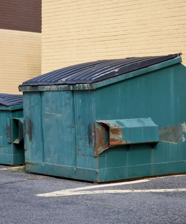 Large commercial green dumpster with a black lid placed behind a yellow brick building.