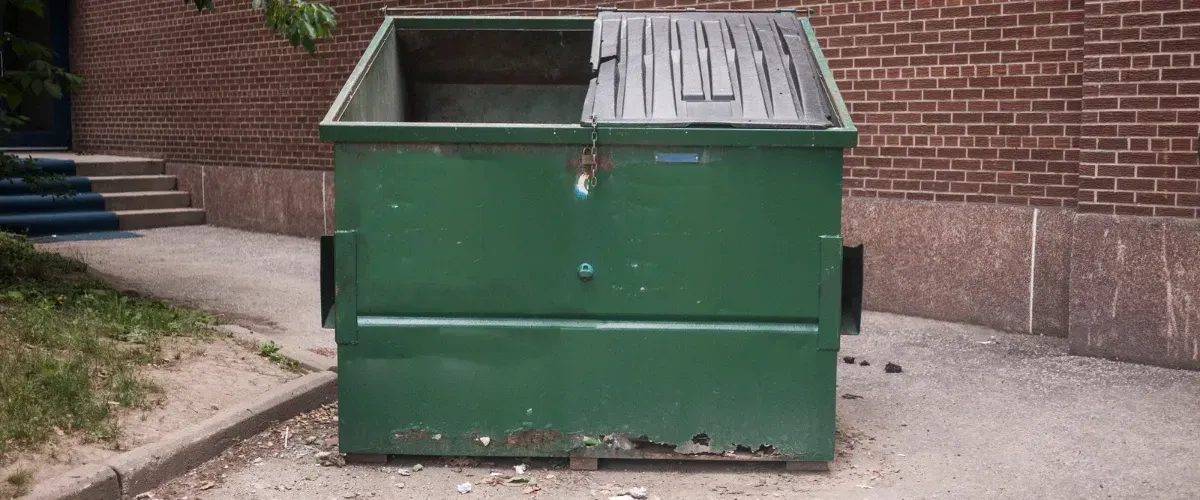 Industrial green dumpster with a partially open lid situated in an alley beside a red brick wall.