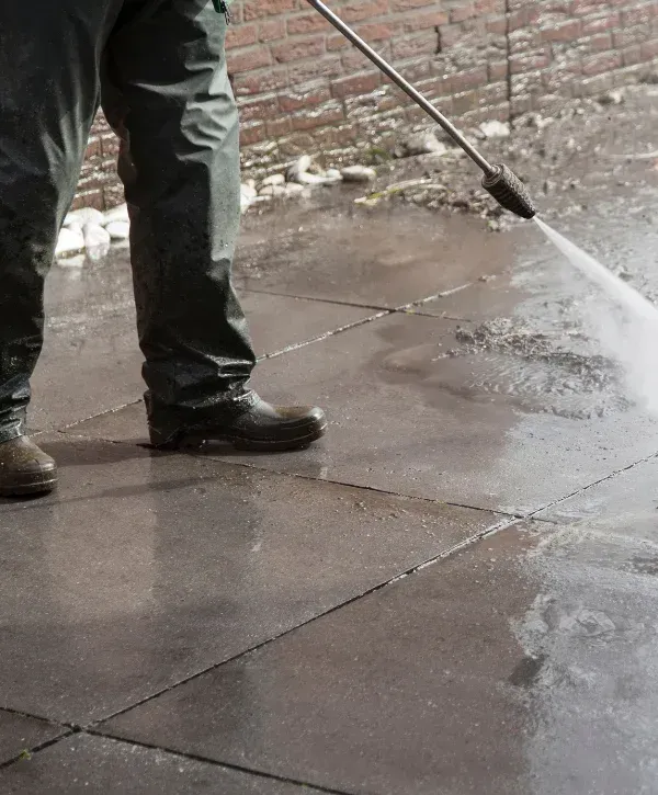 A person in yellow rubber boots power washing a patio made of paving stones next to flowering pink bushes.