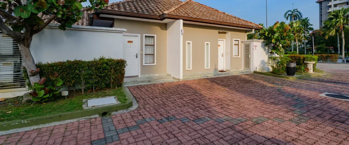 A tan house with a terracotta tile roof and red brick driveway, surrounded by lush green foliage and palm trees.