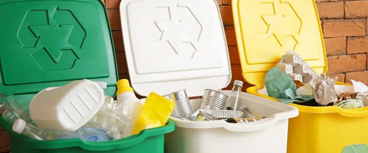 Three color-coded recycling bins, green for plastics, white for metals, and yellow for paper, full against a brick wall.
